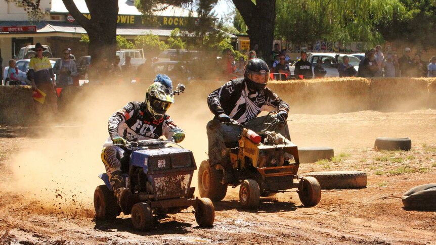 Competitors race around a dirt track on their lawn mowers.
