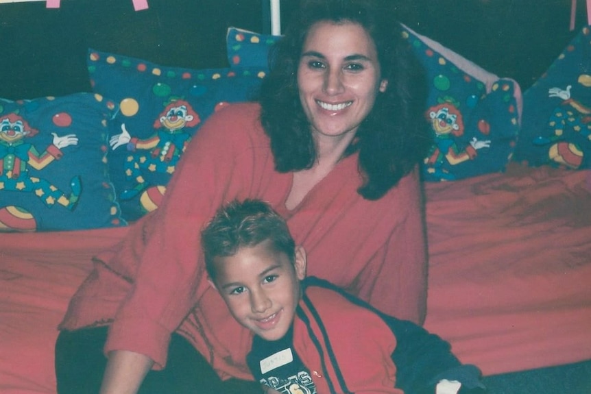 A smiling child with highlights in his hair sits on a bed with his mother. 