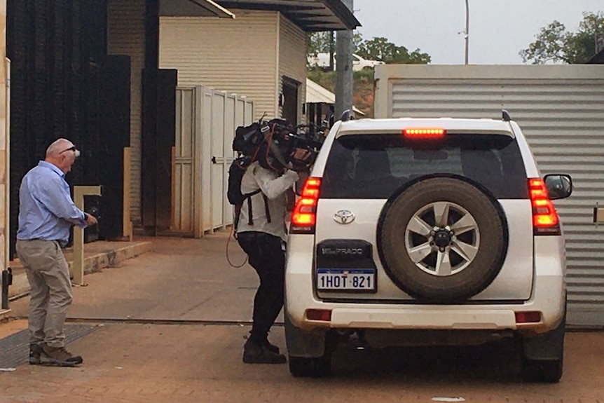 A camera man holding a camera to the window of a parked white car.