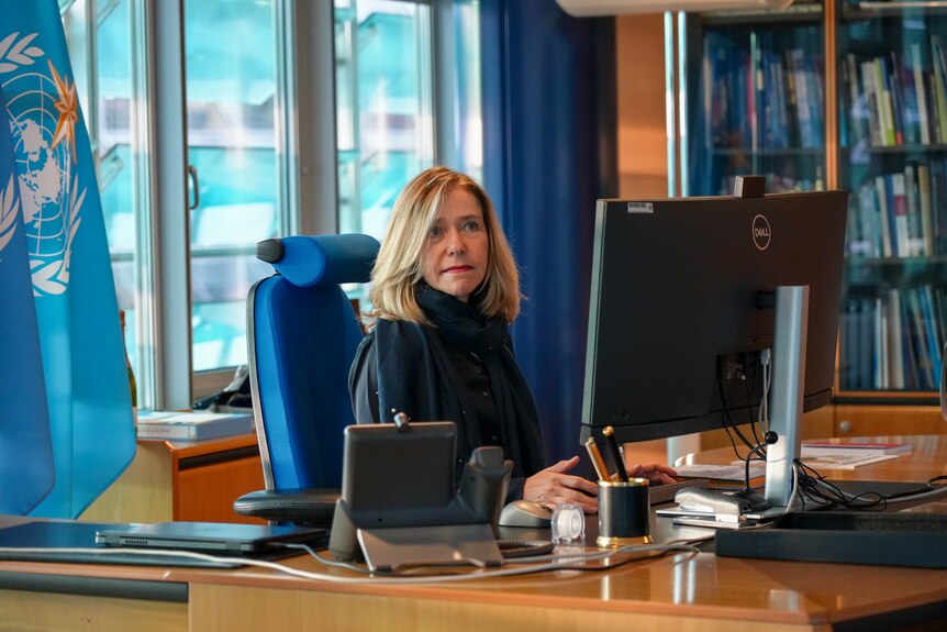 A woman wearing dark clothing, blonde hair and red lipstick sits at a wooden desk in front of a computer, UN blue flag behind.