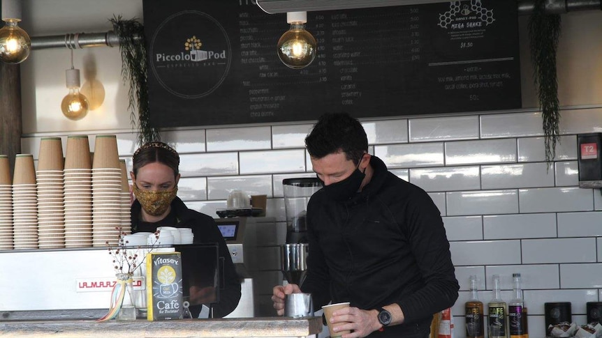 Two male baristas wearing black masks while making coffee.