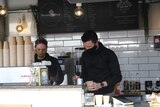 Two male baristas wearing black masks while making coffee.