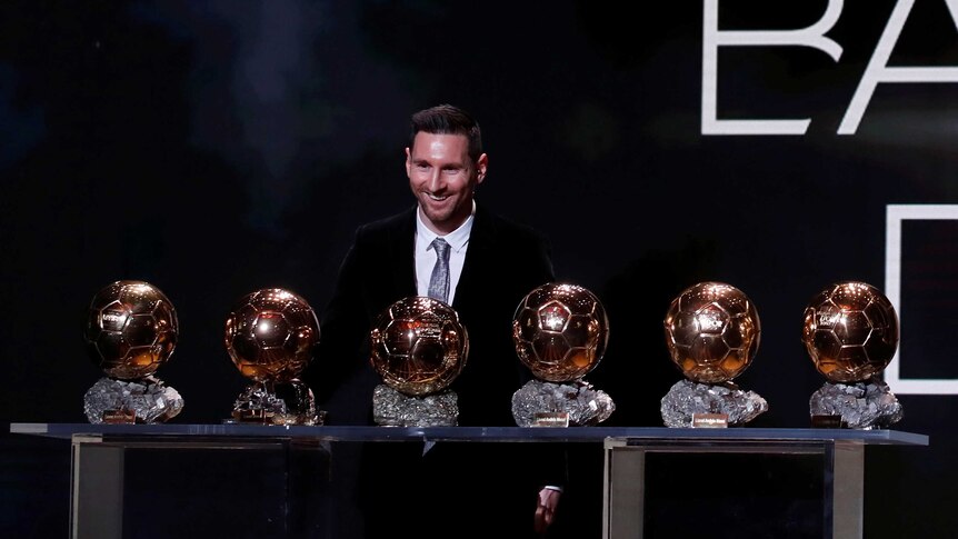 Lionel Messi stands smiling over six Ballon d'Or trophies.