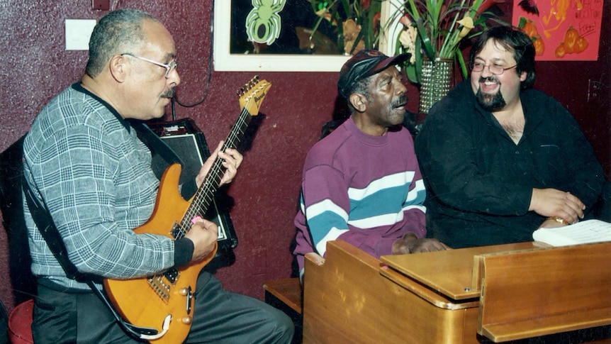 Three male musicians sit side by side; one plays an electric guitar, two others play an organ.