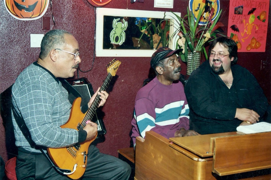 Three male musicians sit side by side; one plays an electric guitar, two others play an organ.