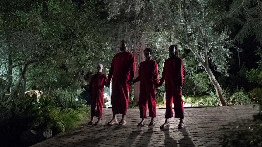 Four imposing figures in red jumpsuits stand on a shadowy drive way, surrounded by thick forest.