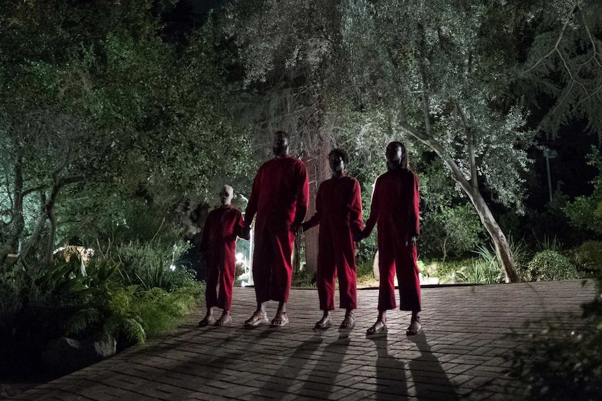 Four imposing figures in red jumpsuits stand on a shadowy drive way, surrounded by thick forest.