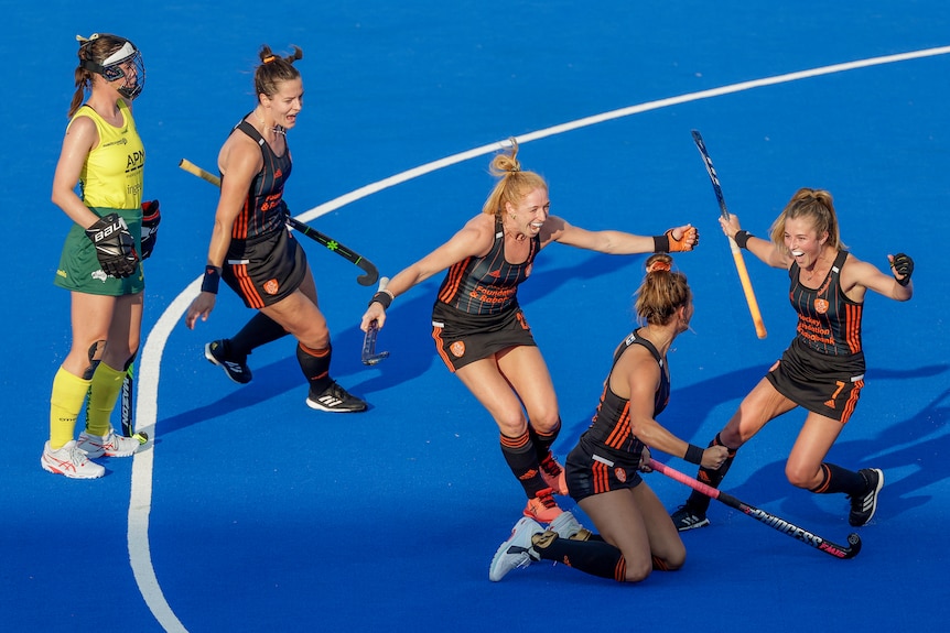 A group of Dutch women's hockey players run towards a teammate who has scored a goal while an Australian stands watching.