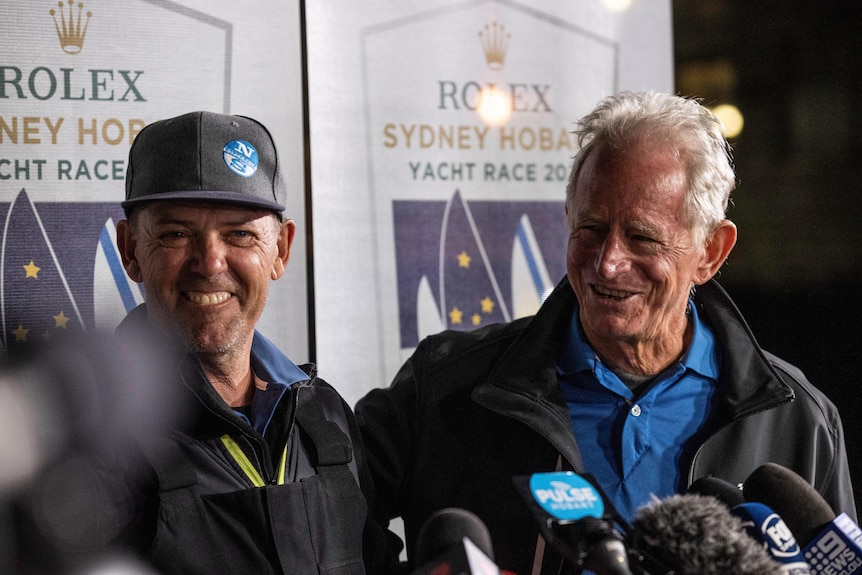Two men smile at a press conference with the Rolex Sydney Hobart Yacht Race banner in the background.