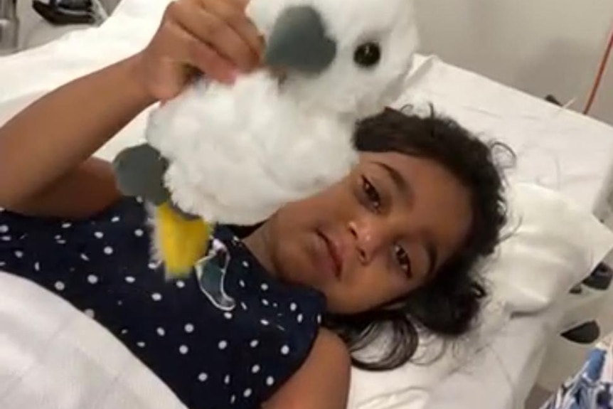 Tharnicaa holds up a stuffed toy while laying in a hospital bed.