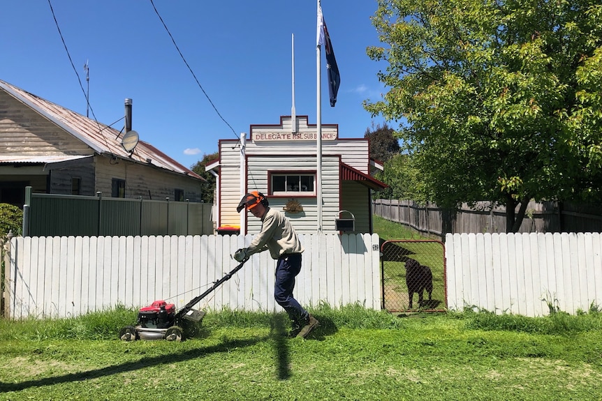 A man mows the lawn out the front of a small hall on a sunny day.