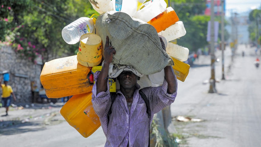 a man in a purple shirt carries multiple empty water bottles and containers above his head 