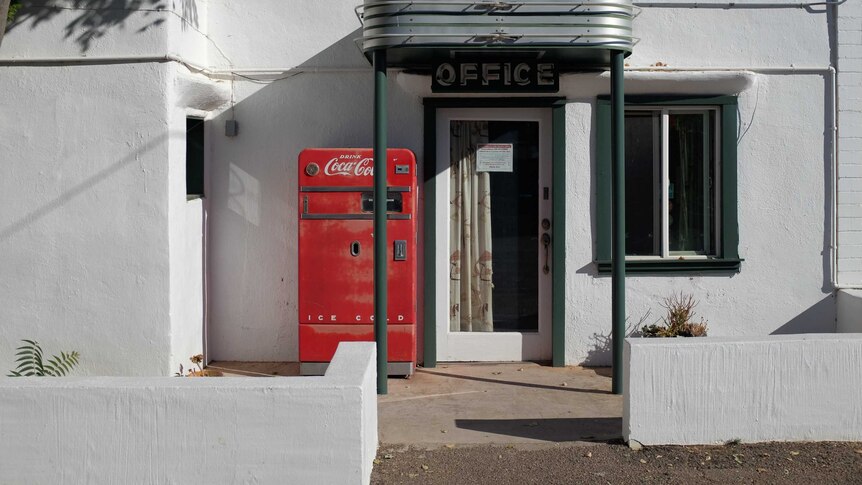 A bright red Coca-Cola vending machine standing in front of a mid-century, low-rise office block in green and white.