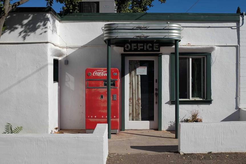 A bright red Coca-Cola vending machine standing in front of a mid-century, low-rise office block in green and white.