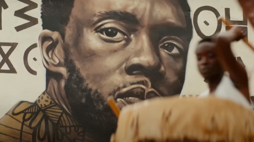 a man plays drums in front of a mural of the Black Panther in a still from the Wakanda Forever trailer
