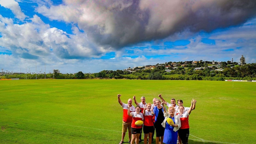 A group stands in the middle of a green football oval raising their thumbs up