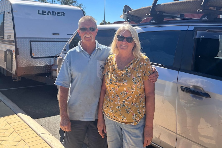 A man in a woman stand arm in arm in front of a four-wheel drive and a caravan. They are smiling.