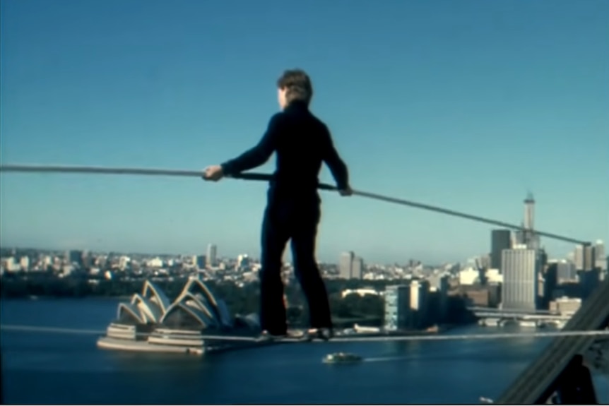 A man in black holds a pole walking on a wire above the harbour and Opera House.