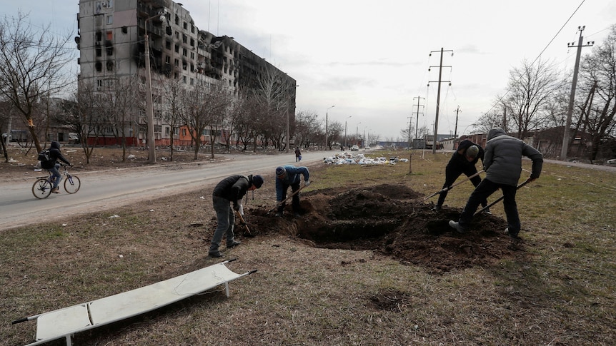 People dig graves in the ground in front of a destroyed building.