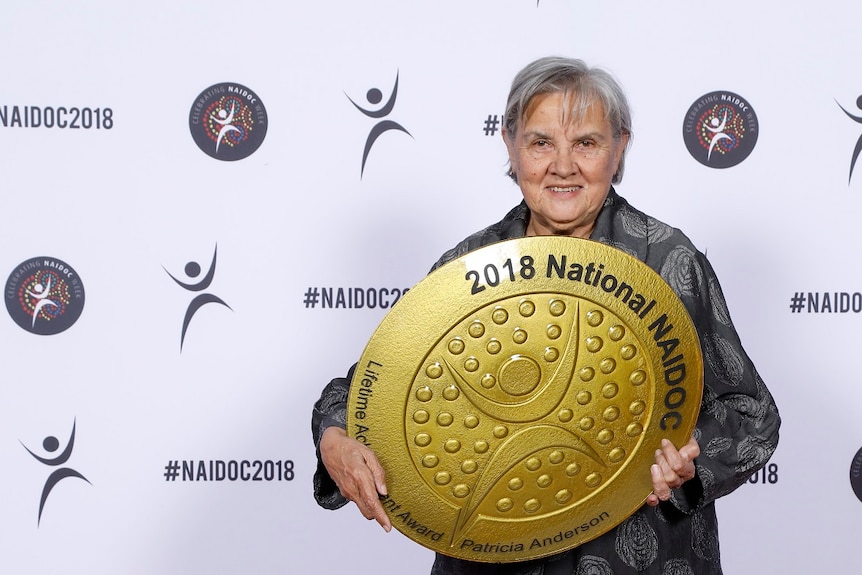 Patricia Anderson in dark coloured evening wear holds a large, round, gold coloured award.