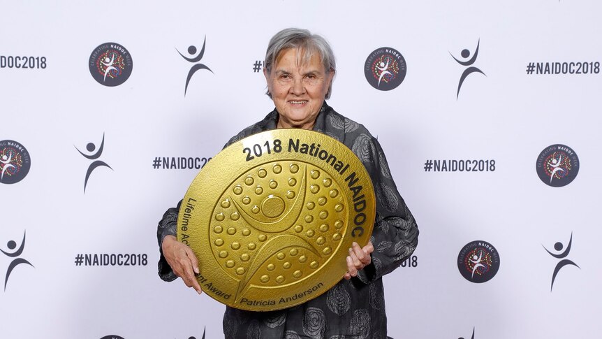 Patricia Anderson in dark coloured evening wear holds a large, round, gold coloured award.