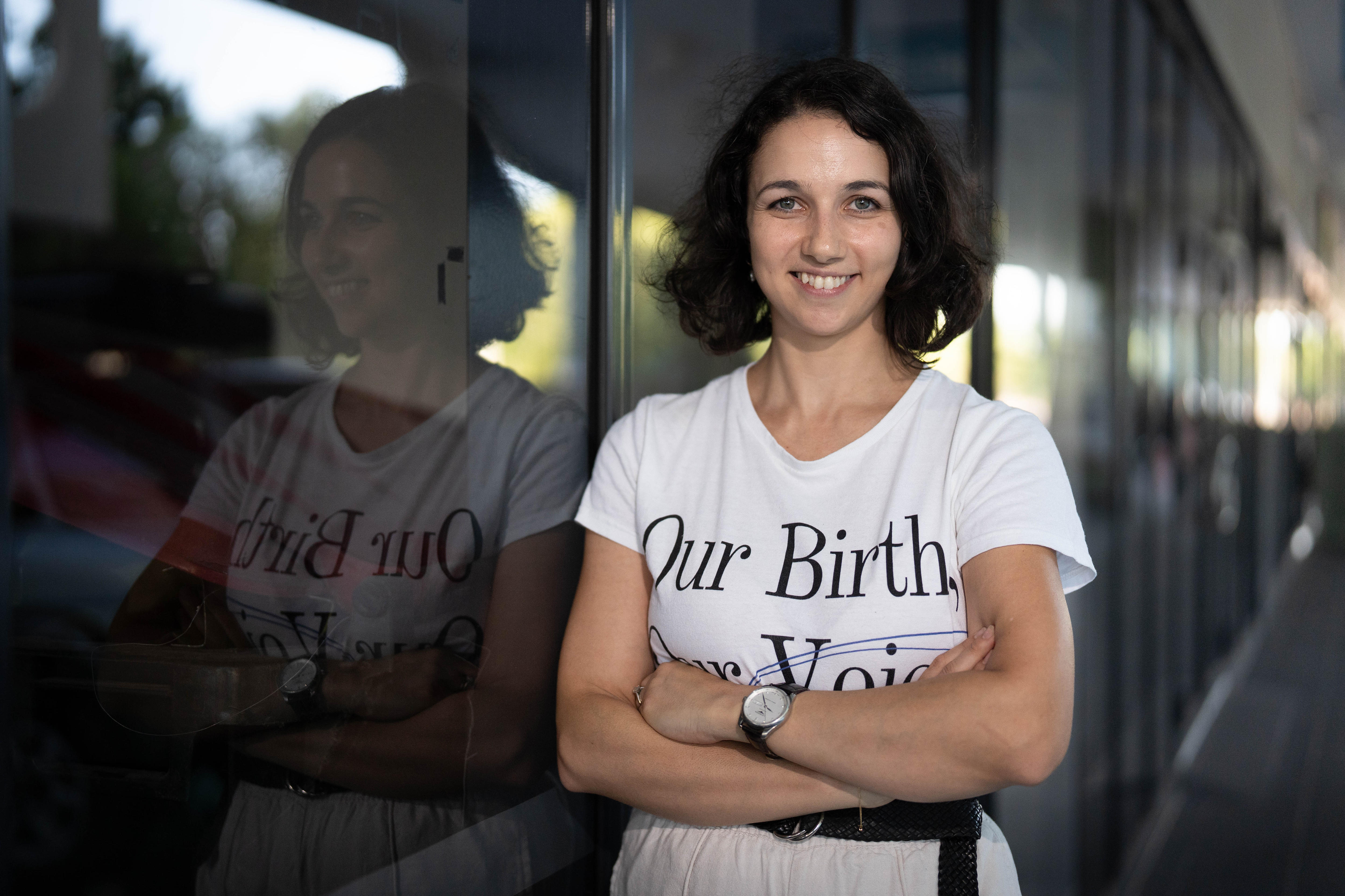 A young woman, Danielle Muhall, wearing a Our Birth Our Voice shirt smiles at the camera.