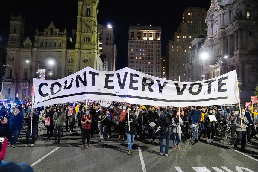 A big group of people in masks march through a city square at night with 'count every vote' sign.