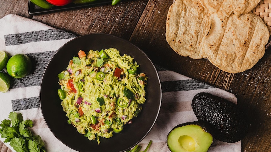 Overhead shot of a bowl of guacamole next to a cut avocado, tortillas, a lime and chillies set on a tablecloth on a wooden table