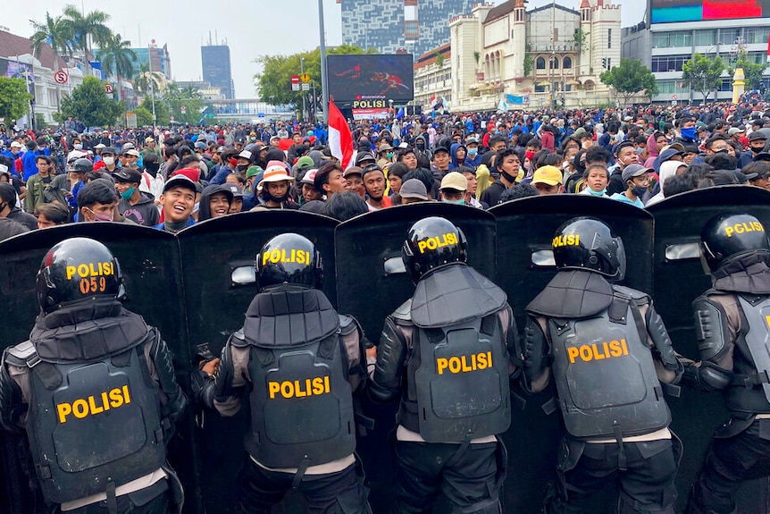 Black-clad riot police form a barricade against a sea of protesters who billow out in a large square amid Jakarta's buildings.