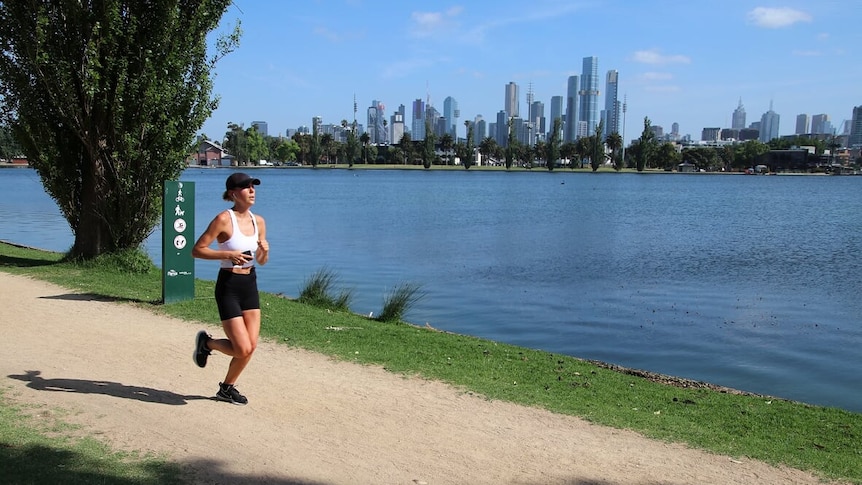 A woman jogs along a path with Melbourne's city skyline in the background.