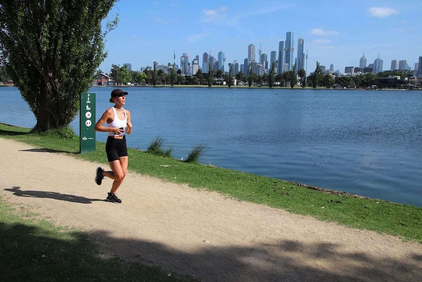 A woman jogs along a path with Melbourne's city skyline in the background.