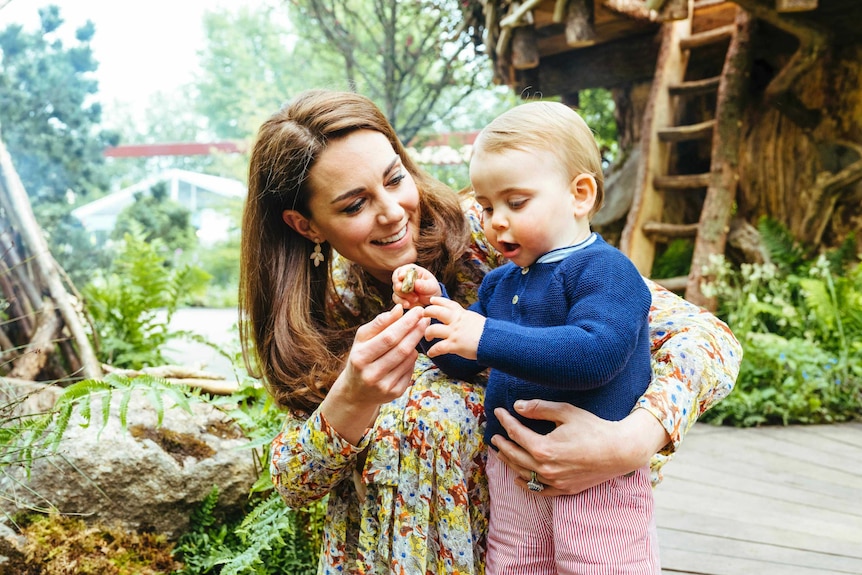 The Duchess of Cambridge looks at her son Prince Louis as he holds a flower