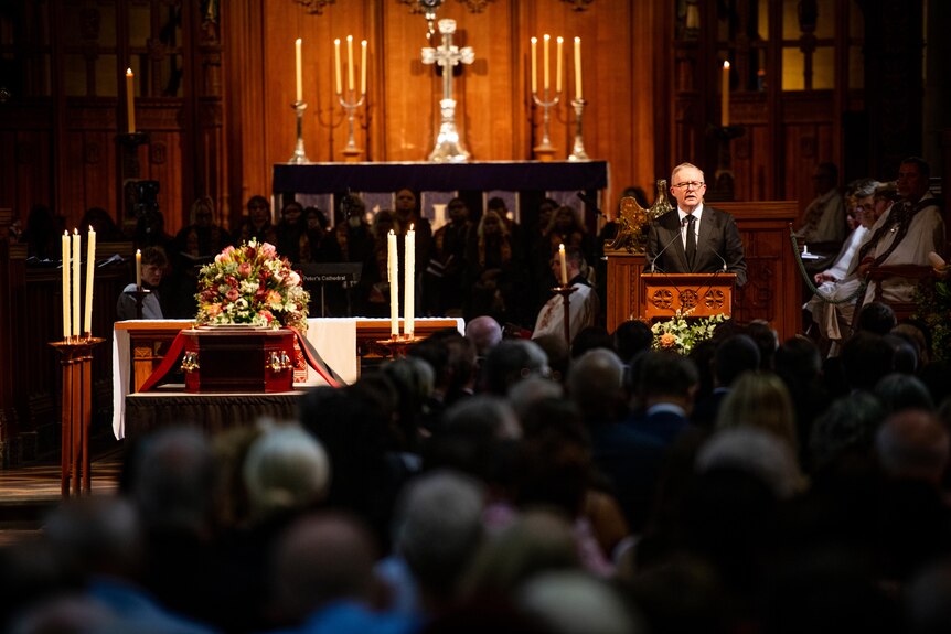 Anthony Albanese speaks at a lecturn next to a coffin adorned with flowers in front of mourner inside a cathedral