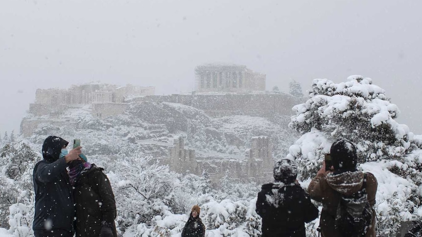People take pictures in front of the ancient Acropolis hill during a snowfall.
