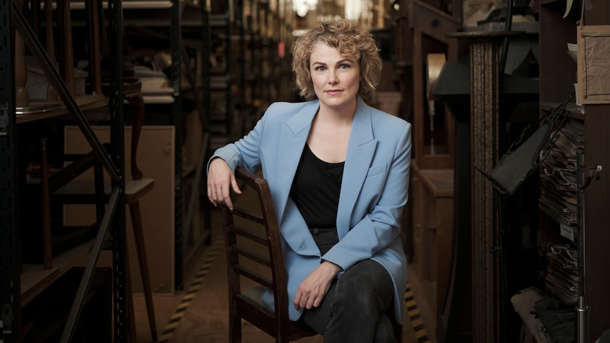 A blonde, curly-haired woman sits on a chair wearing a blue blazer, backstage at a theatre company