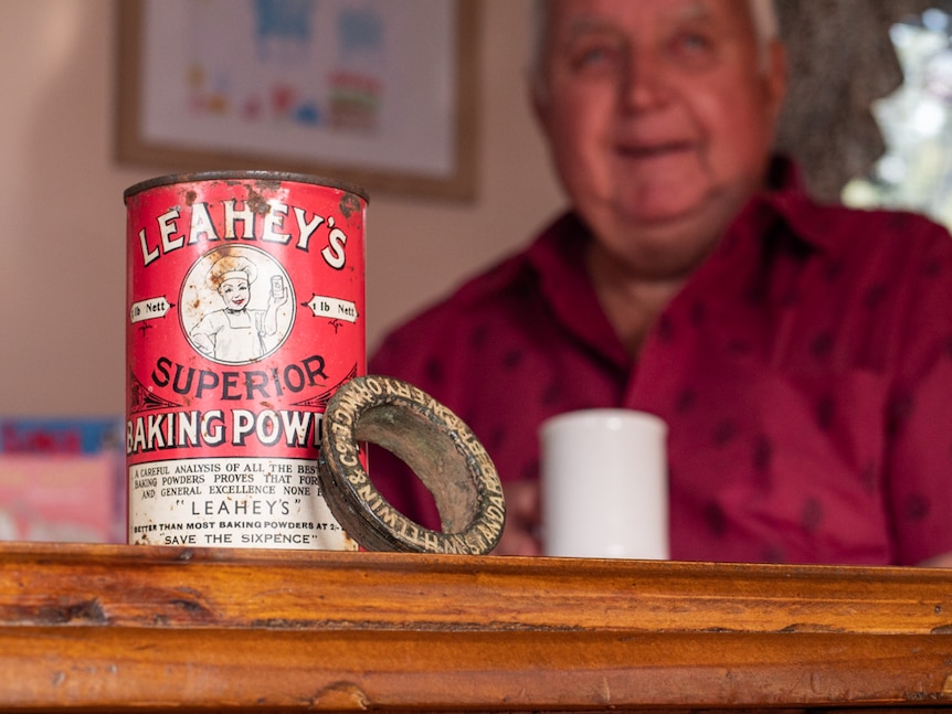 A Leahey's baking powder tin and a beer bung in the foreground with an older man sitting with a mug.