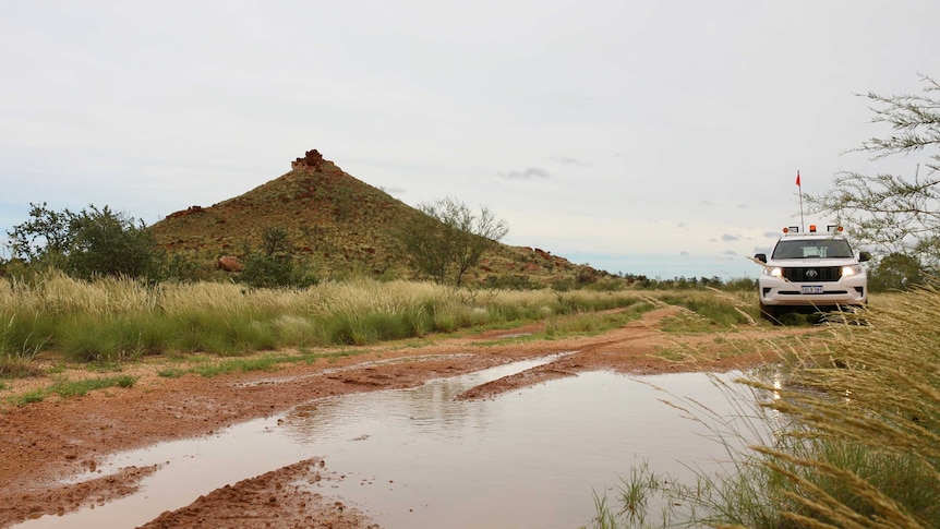 A waterlogged dirt road with a white four-wheel-drive alongside it and a hill in the background.
