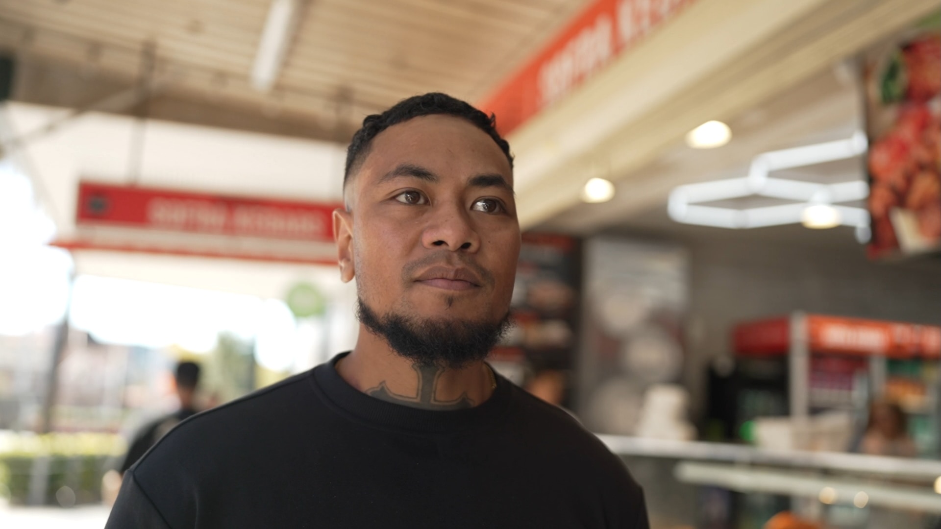 A man in a black t-shirt walking down a street. A partial tattoo is visible on his neck.