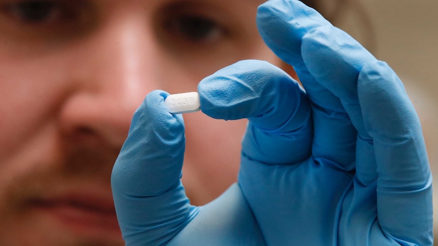 A man wearing blue gloves holds a white pill up to the camera.