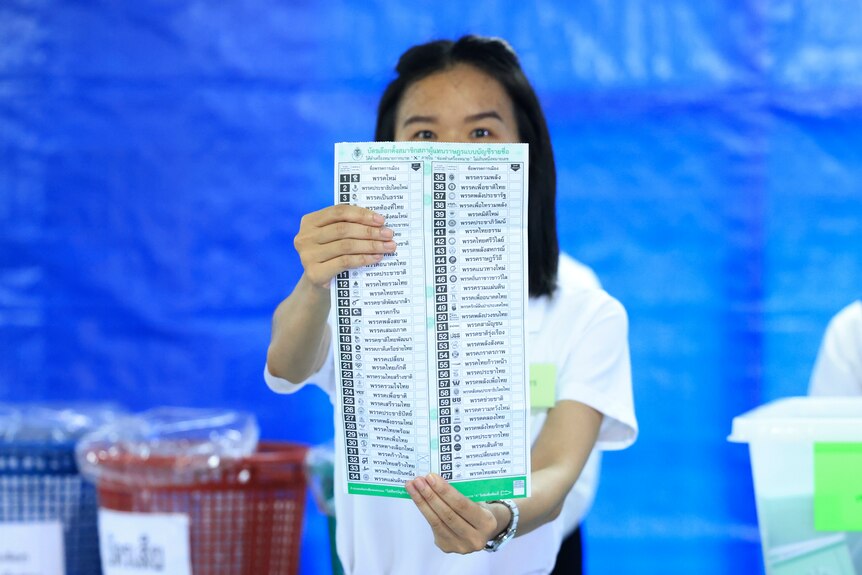 A Thai woman standing in front of a blue backdrop, holds up a large voting ballot paper.
