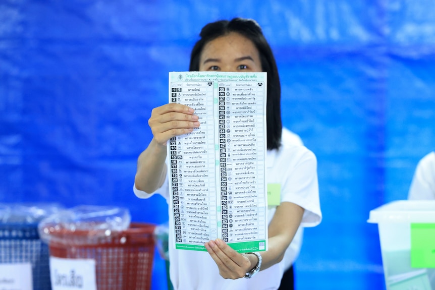 A Thai woman standing in front of a blue backdrop, holds up a large voting ballot paper.