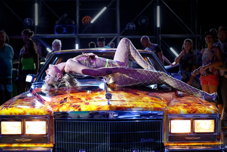 A woman in fishnets and gold bikini strikes a pose on the bonnet of a car painted with flames