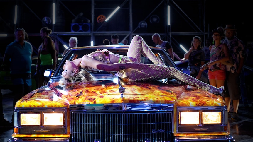 A woman in fishnets and gold bikini strikes a pose on the bonnet of a car painted with flames