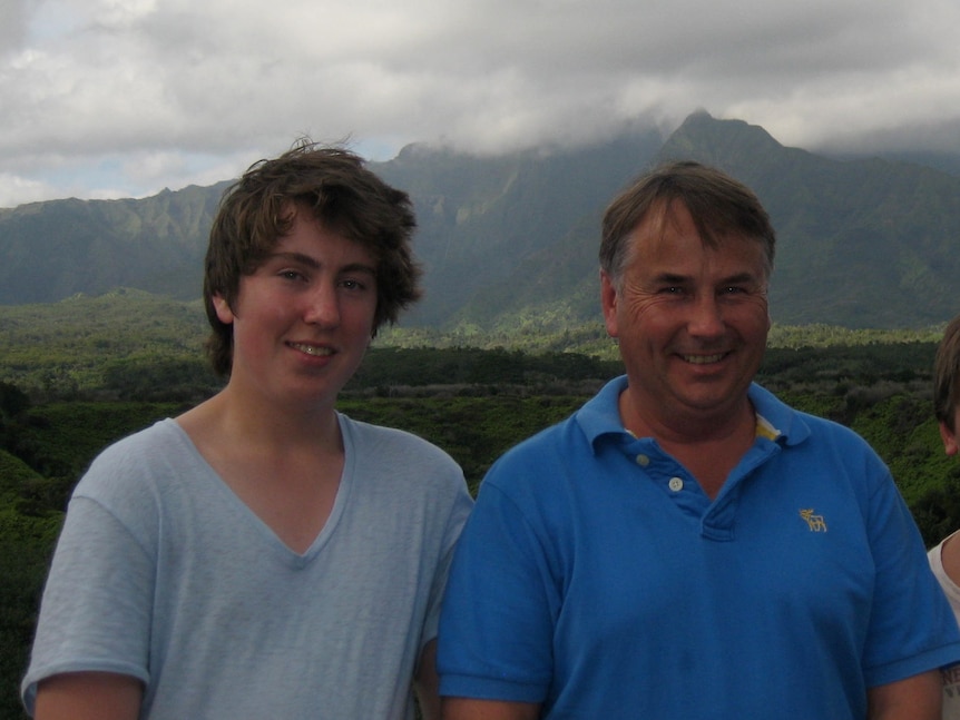 A father and two sons smile in front of a mountain.