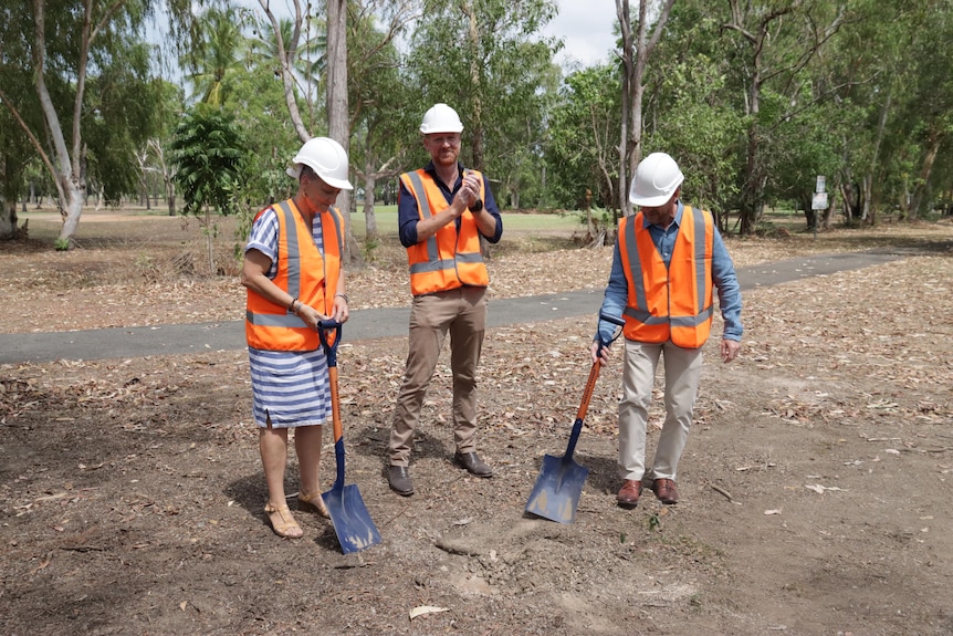 Three people wearing hardhats and bright construction vests put shovels in the ground.