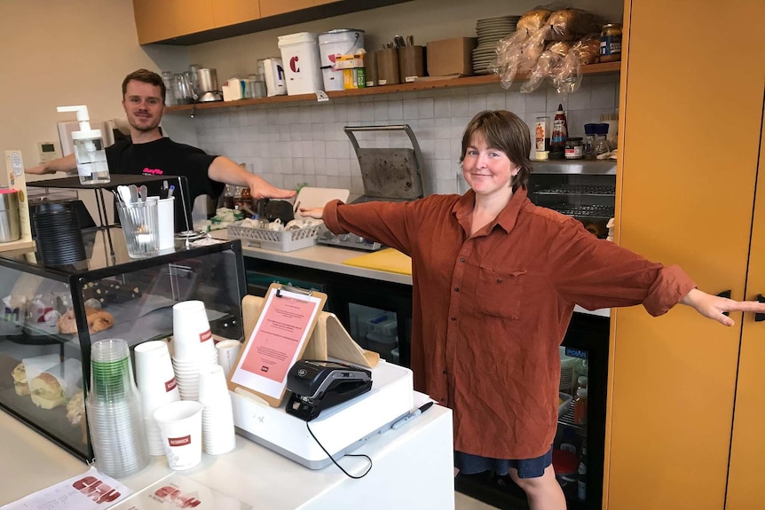 Two people hold their arms apart from each other, standing behind a cafe counter and smiling.