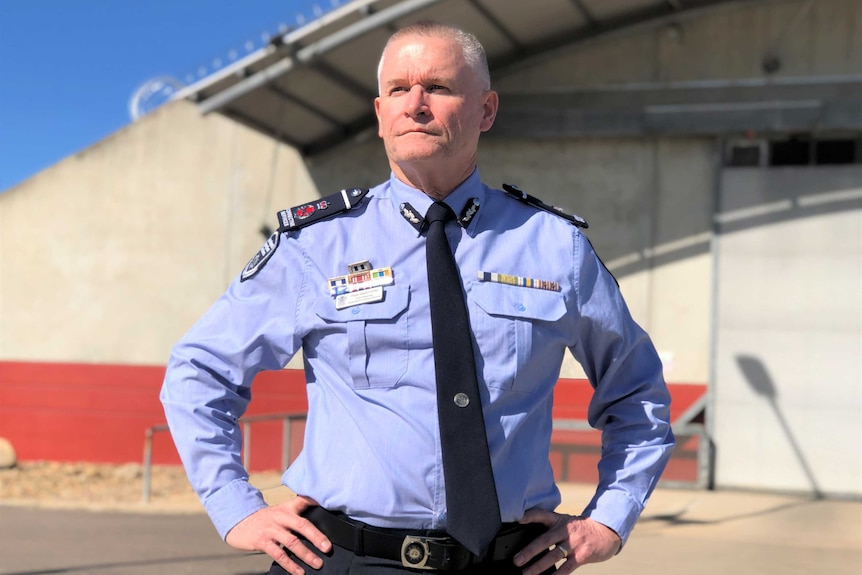 A man wearing a corrective services uniform stands outside a prison with his hands on his hips