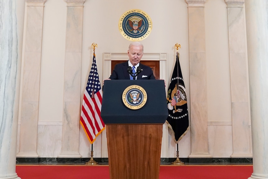 President Joe Biden, dressed in a suit, speaks from behind a lectern, flanked by two flags and a United States crest