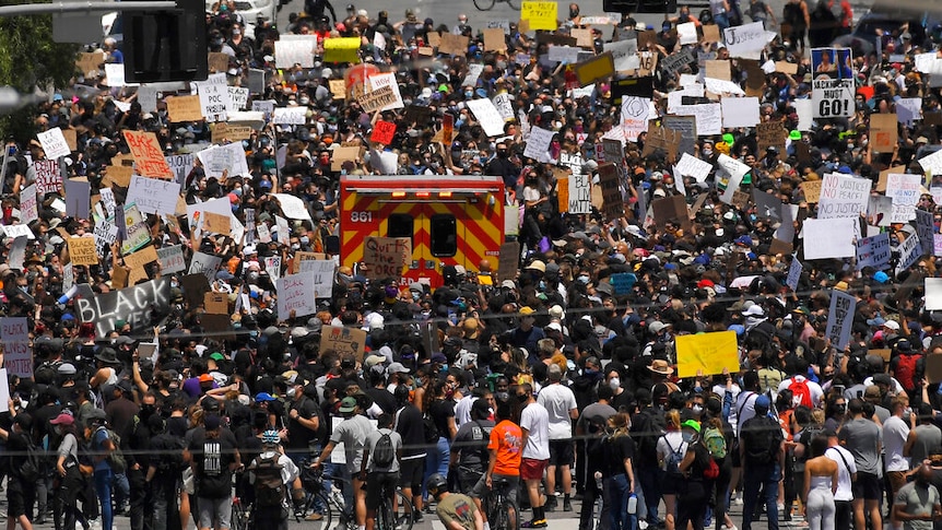 A crowd of hundreds holding signs surround an emergency vehicle.