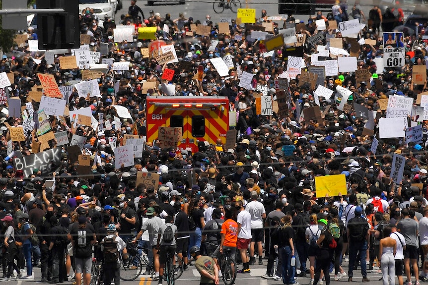 A crowd of hundreds holding signs surround an emergency vehicle.